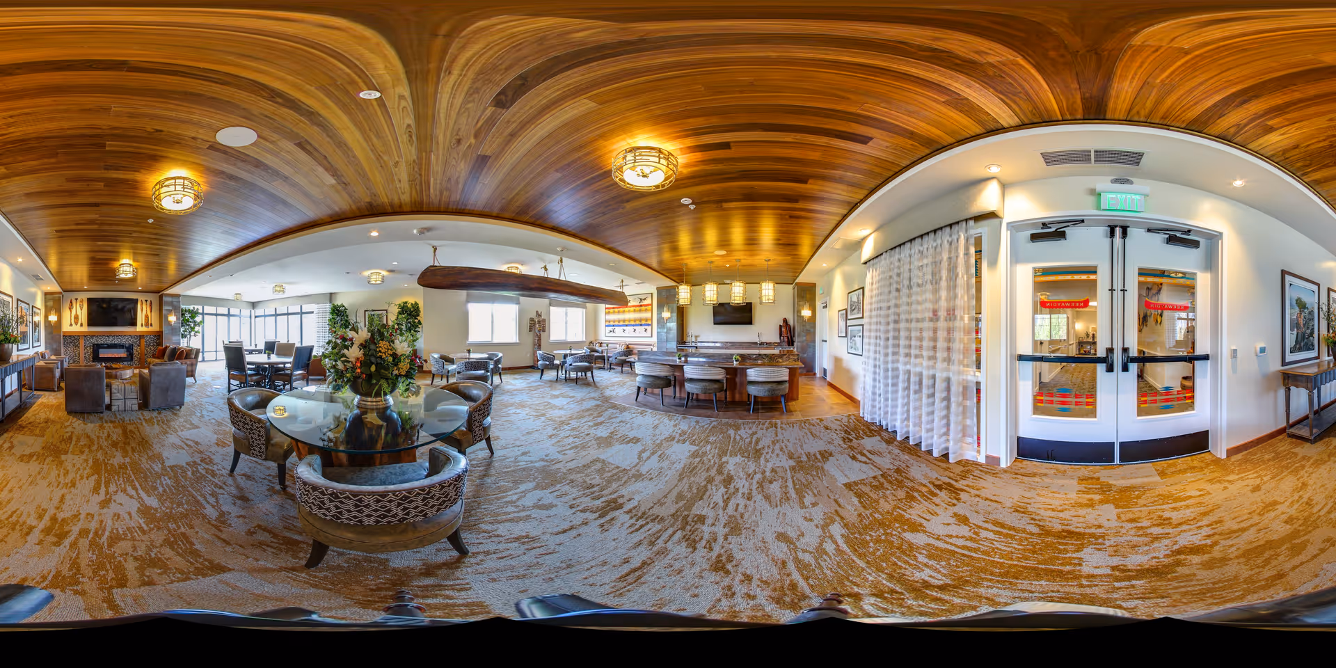 A spacious and well-lit living room area in a senior living facility with a wooden ceiling, carpeted floor, and multiple seating arrangements including armchairs and round tables. There is a fireplace with a TV mounted above it on one side, large windows letting in natural light, and decorative plants and artwork on the walls. The entrance doors with exit signs are visible on the right side.
