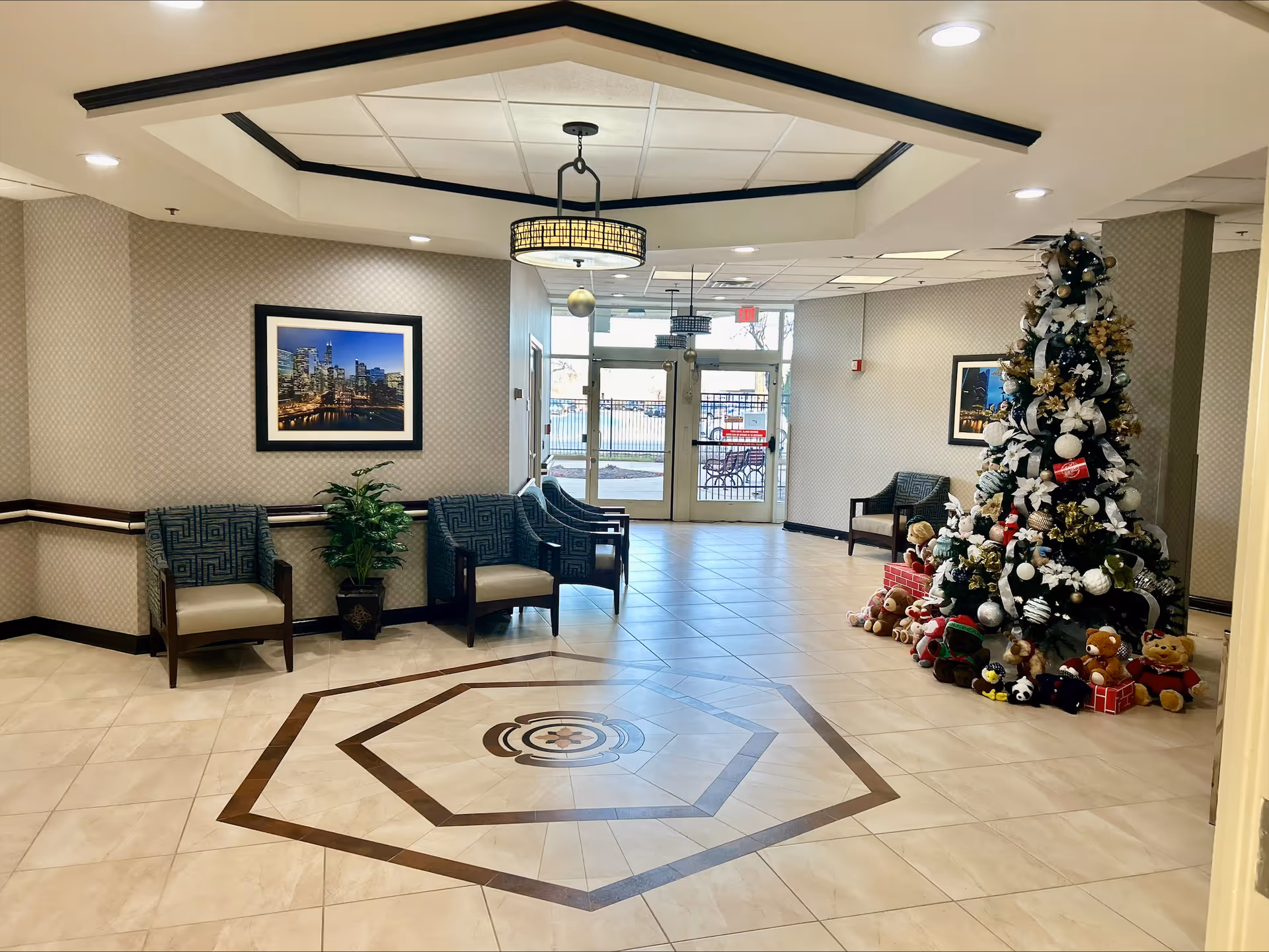 A spacious lobby area with tiled floors featuring a geometric design in the center. There are several cushioned chairs with patterned upholstery along the walls, a potted plant, and framed cityscape photographs hanging on the walls. A decorated Christmas tree with ornaments, ribbons, and stuffed animals at its base is positioned on the right side near the entrance doors. The ceiling has recessed lighting and a decorative light fixture.