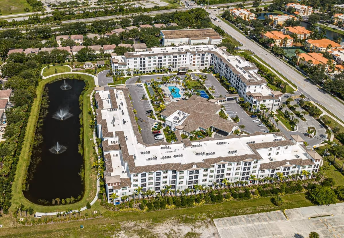 Aerial view of a large senior living facility named Arcadia Gardens, featuring a multi-story building arranged in a U-shape around a central courtyard with a swimming pool, palm trees, and parking areas. To the left of the building is a long pond with three water fountains, surrounded by greenery and walking paths. The facility is located near a main road and residential neighborhoods.