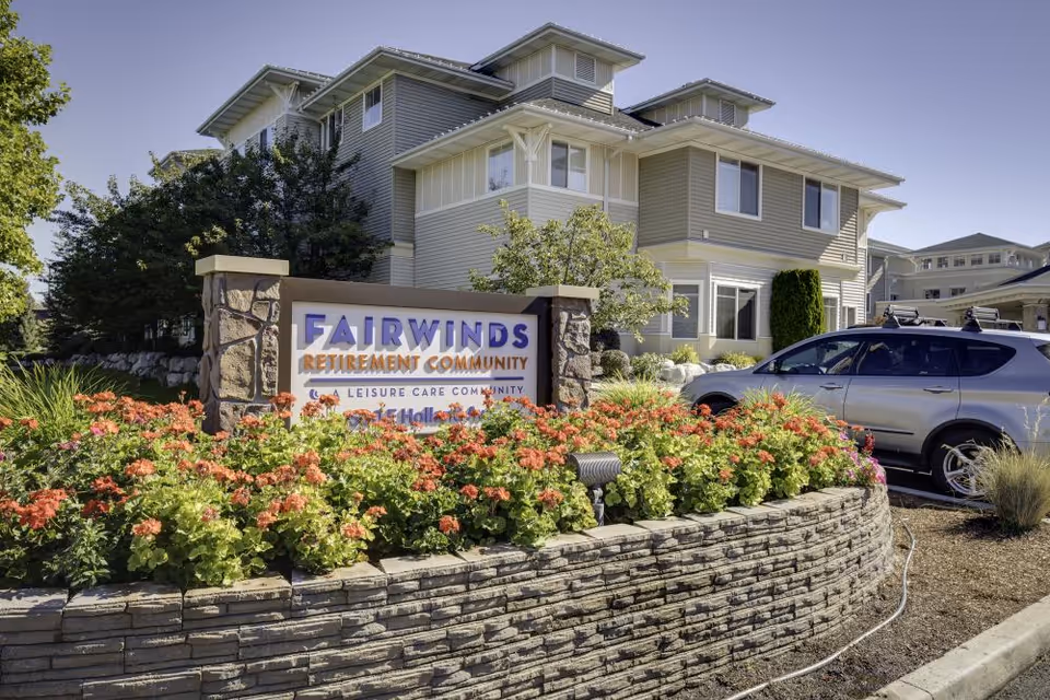 Exterior view of Fairwinds Retirement Community building with a stone and flower bed in front displaying the community's sign. A silver SUV is parked nearby under a clear sky.
