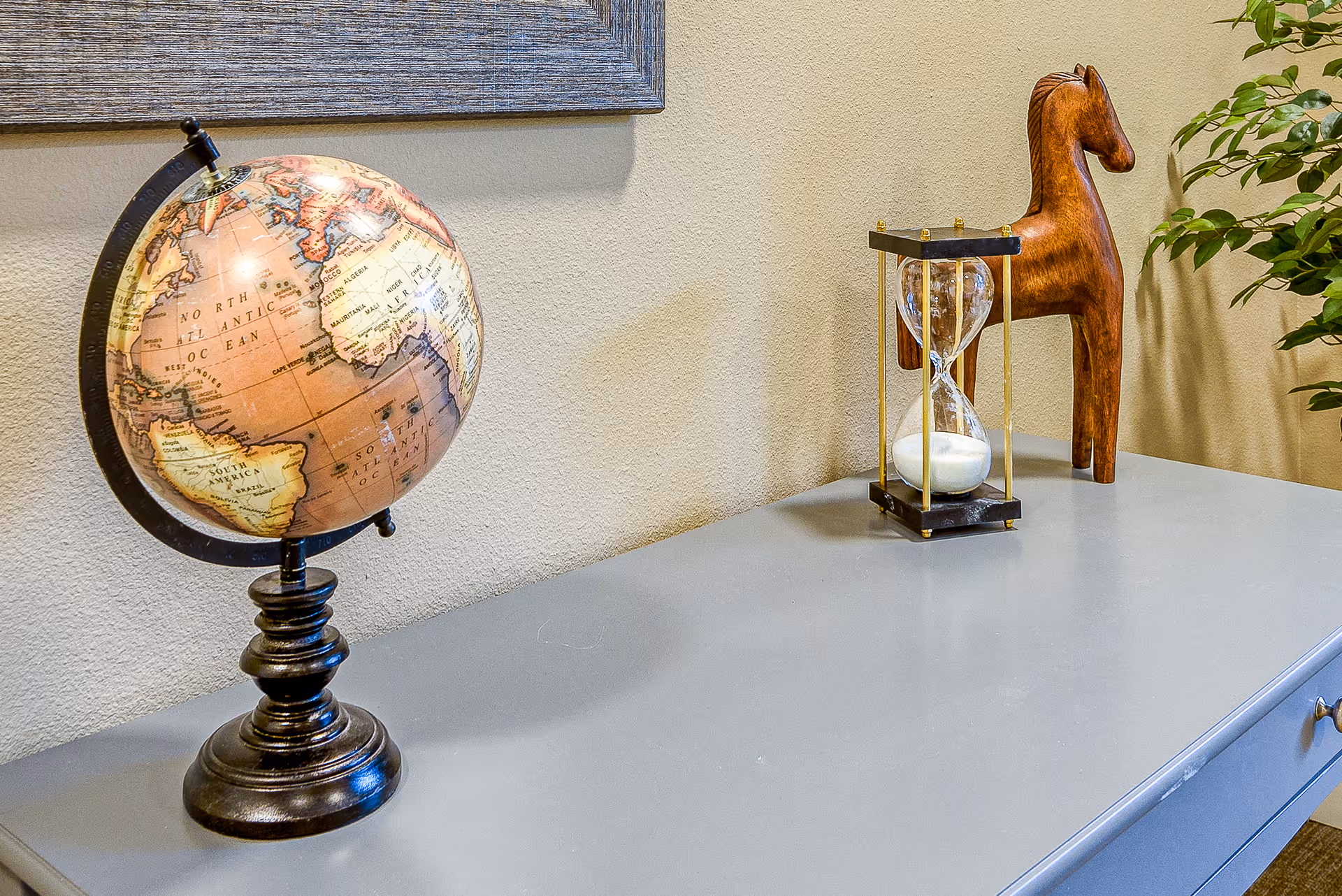 A decorative globe, hourglass, and wooden horse figurine arranged on a gray console table against a beige wall.