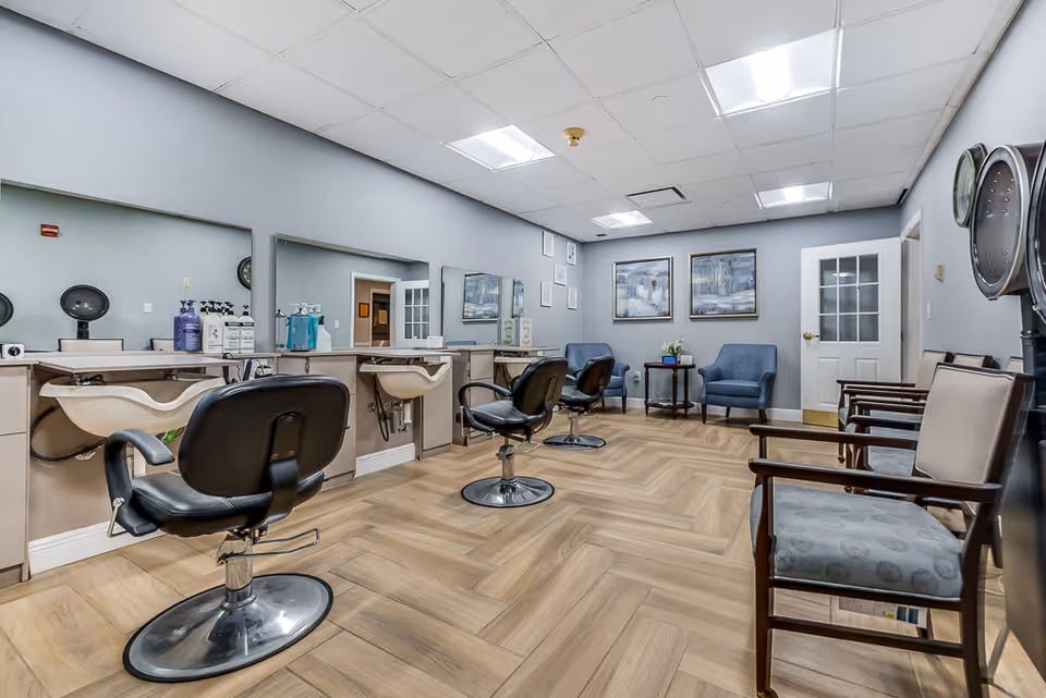 Interior view of a senior living facility's hair salon with three black salon chairs in front of sinks and mirrors. The room has light wood flooring, gray walls, framed artwork, and a seating area with blue armchairs and wooden chairs along the wall.