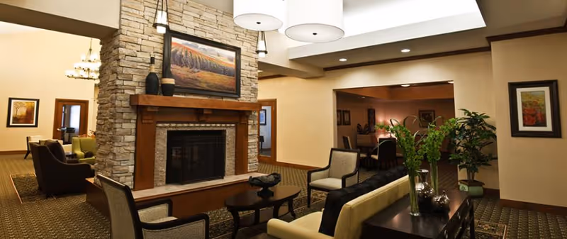 A cozy living room area featuring a stone fireplace with a wooden mantel, a framed landscape painting above it, and comfortable seating including armchairs and a sofa. The room is warmly lit with ceiling lights and a chandelier visible in the background. There are decorative plants and framed artwork on the walls, creating a welcoming atmosphere.
