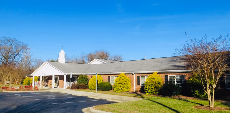 Exterior view of a single-story brick building with a gray roof and white trim, surrounded by neatly trimmed bushes and trees under a clear blue sky. There is a paved parking area in front with marked parking spaces.