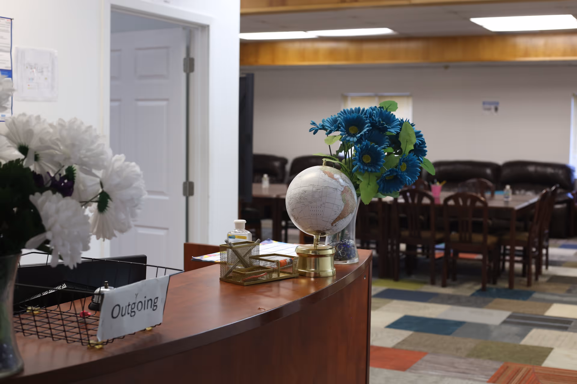 Interior view of a senior living facility reception area with a curved wooden desk. On the desk, there is a globe, a vase with blue flowers, a wire basket labeled 'Outgoing', and a bottle of hand sanitizer. In the background, there are tables and chairs with dark brown leather couches along the wall, and a colorful patterned carpet on the floor.
