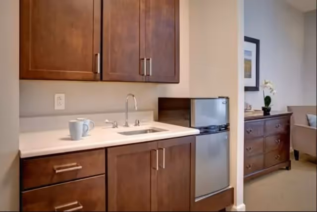 A small kitchenette area with wooden cabinets, a white countertop, a sink with a faucet, and a white mug on the counter. To the right, there is a compact stainless steel refrigerator. In the background, part of a living area is visible with a wooden dresser, a framed picture on the wall, a potted plant, and a beige armchair.