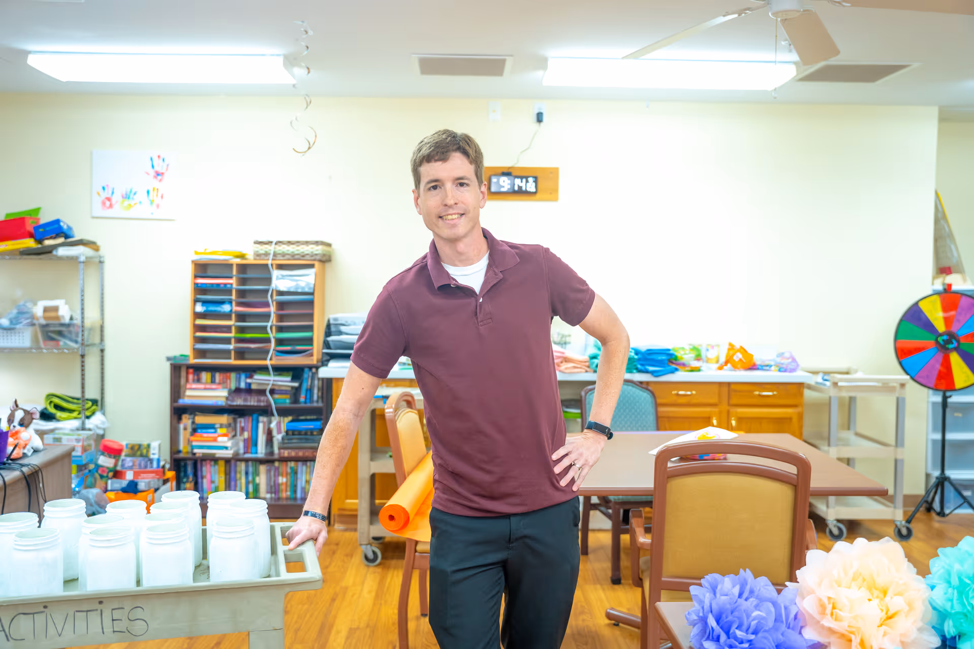 A man in a maroon polo shirt and black pants stands smiling in a brightly lit activity room. The room contains tables, chairs, shelves with books and craft supplies, colorful paper flowers, and a colorful spinning wheel. The floor is wooden, and the walls are light-colored.