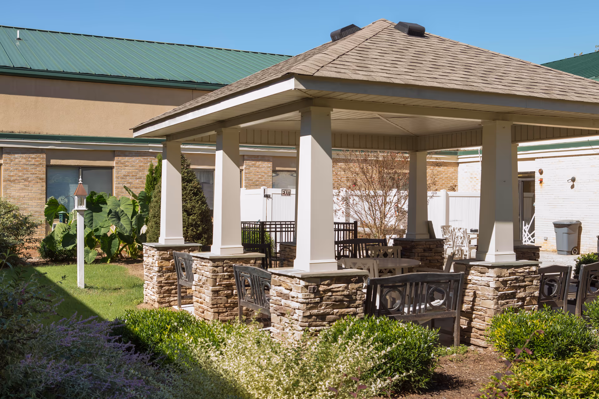 Outdoor covered seating area with stone pillars and a shingled roof surrounded by greenery and bushes, located in the courtyard of a building with a green metal roof and beige walls.