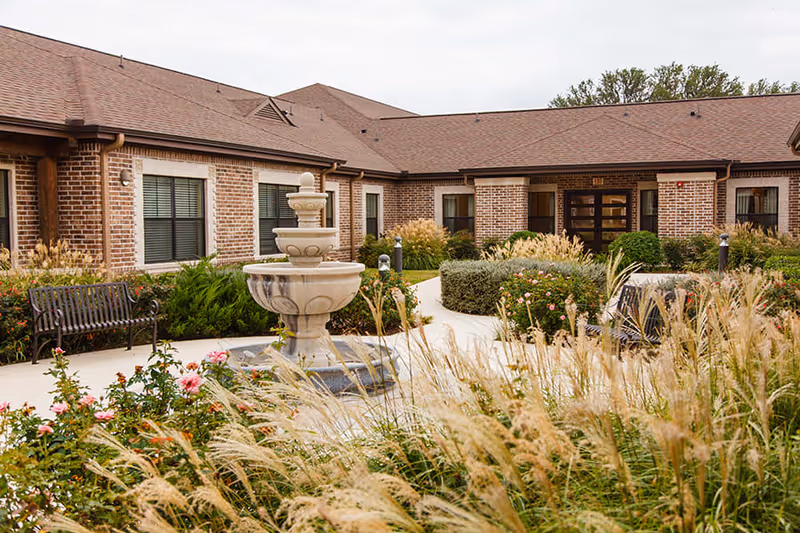 Outdoor courtyard area of a senior living facility featuring a central stone fountain surrounded by landscaped bushes, flowers, and ornamental grasses. The courtyard is bordered by a brick building with multiple windows and a bench along the walkway.