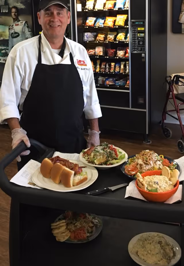 A man wearing a white chef's coat, black apron, and black cap stands behind a food service cart with plates of food including a hot dog, salad, and other dishes. Behind him is a vending machine and a walker is visible to the right.