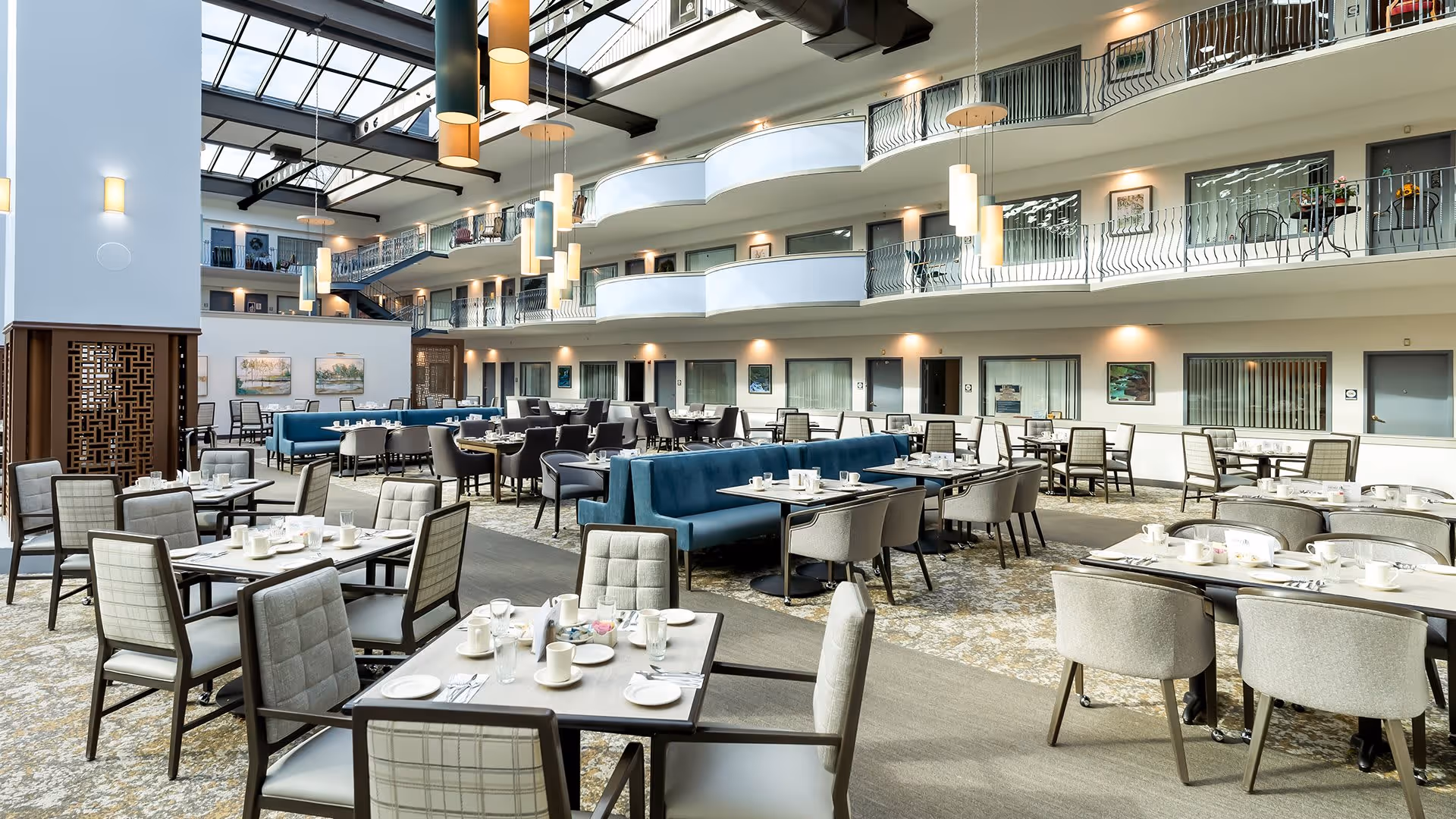 Spacious and well-lit dining area in a senior living facility with multiple tables and chairs arranged neatly. The room features a high ceiling with large skylights, modern pendant lights, and a second-floor balcony overlooking the dining space. Tables are set with white cups, plates, and utensils, ready for a meal.