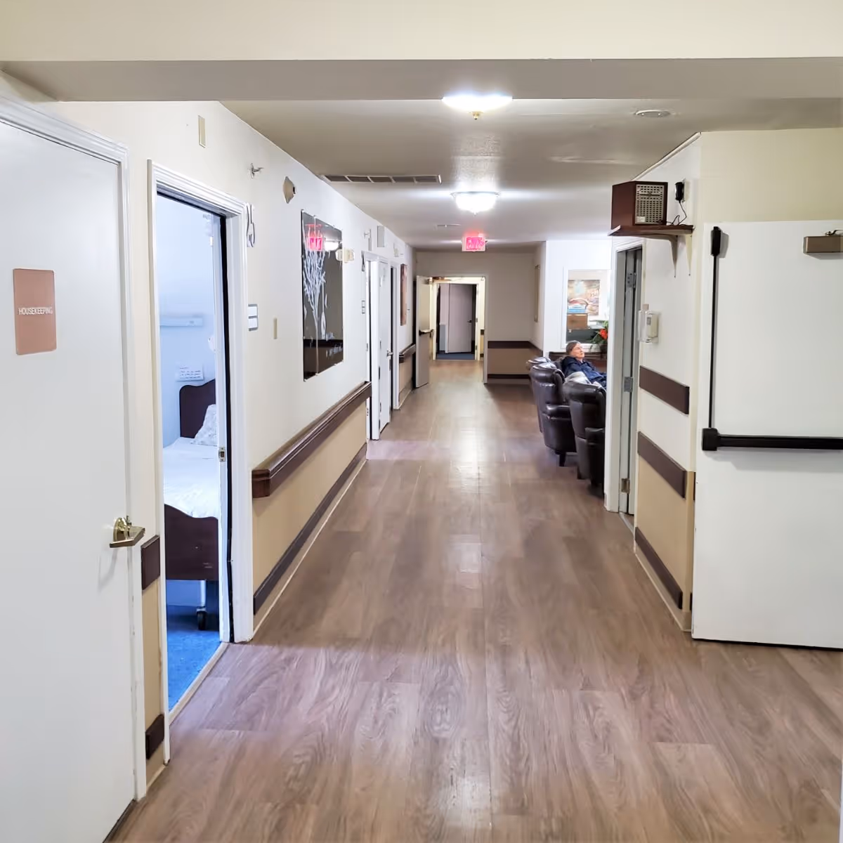 A long hallway in a senior living facility with wood-patterned flooring and beige walls. On the left, there are doors including one labeled 'HOUSEKEEPING' and an open door showing a bedroom with a bed. On the right, there is a seating area with a person sitting on a dark leather chair. The hallway is well-lit with ceiling lights and has handrails along the walls.