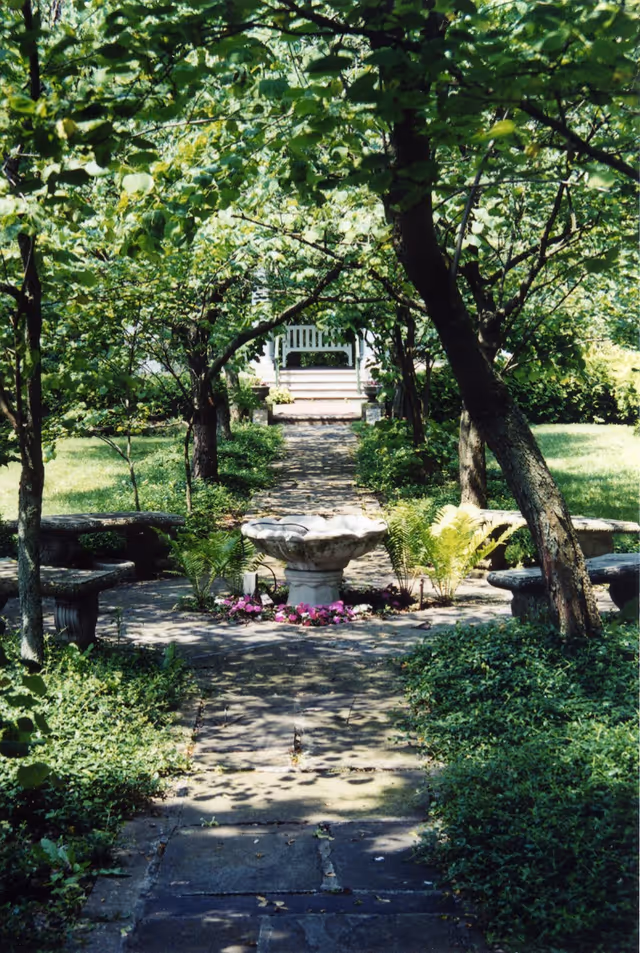 Shaded garden pathway lined with trees leading to a stone fountain and benches with a white porch bench visible in the background.