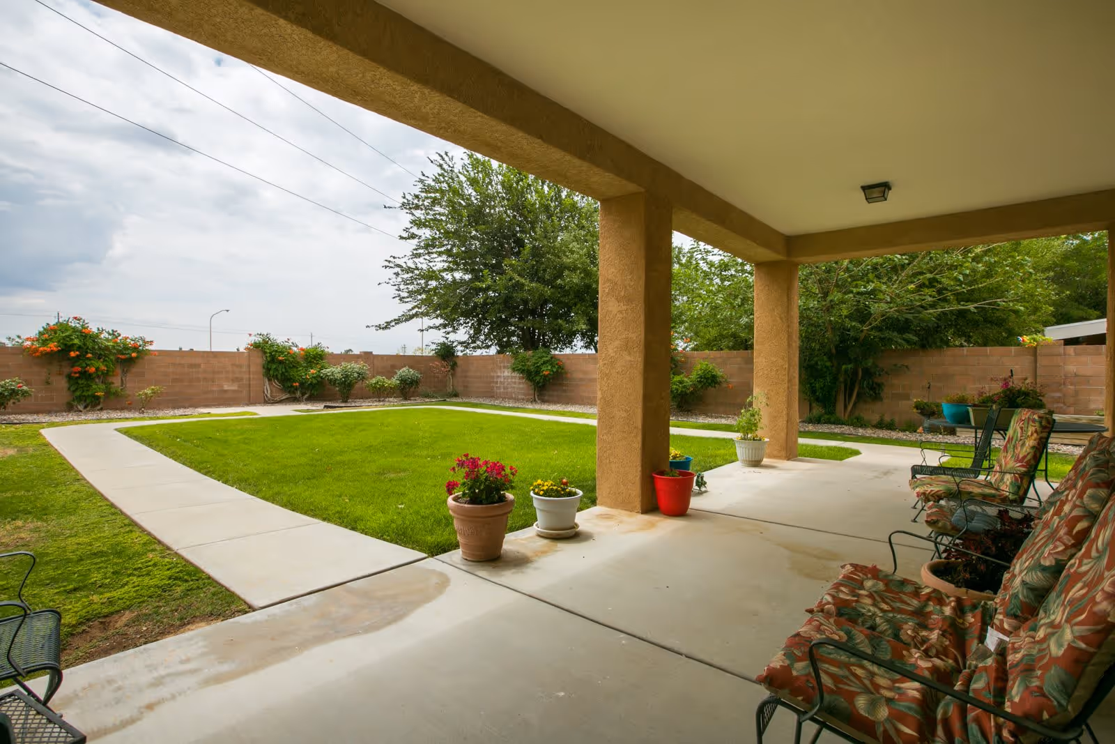 Covered patio area with cushioned chairs and potted plants overlooking a green lawn and garden with a brick wall and trees in the background under a cloudy sky.