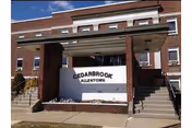 Exterior view of Cedarbrook Senior Care and Rehab building with a brick facade and a large sign in front reading 'CEDARBROOK ALLENTOWN'. There are stairs on both sides of the sign leading up to the building entrance.