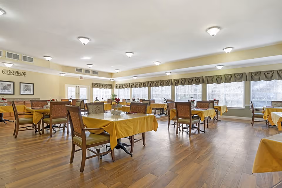 A spacious dining room with multiple tables covered in yellow tablecloths, each surrounded by wooden chairs with cushioned seats. The room has large windows with valance curtains allowing natural light to fill the space. The floor is wooden, and the ceiling has several light fixtures. There are framed pictures on the wall and small flower arrangements on each table.
