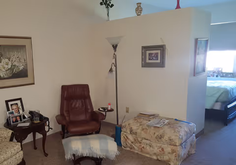 A cozy living area with a brown leather recliner chair, a small side table with framed photos, a floral patterned ottoman with papers on top, a floor lamp, and a partial view of a bedroom with a bed and window in the background.