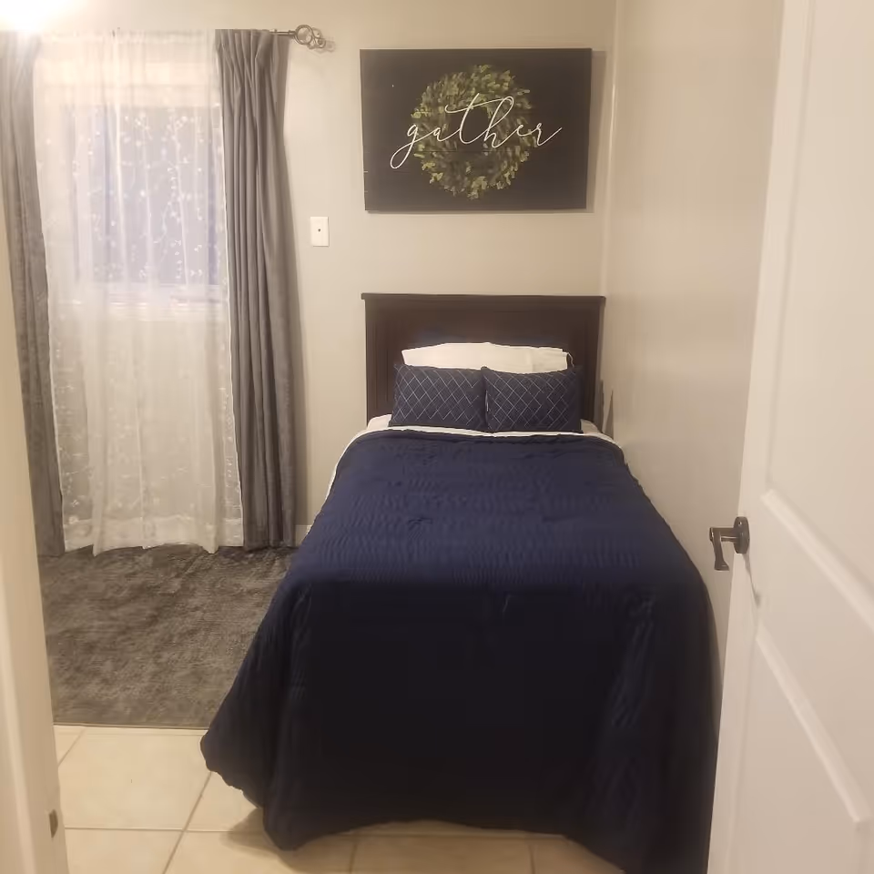 A small bedroom with a single bed covered in a dark blue quilt and two matching pillows. Above the bed is a wall decoration featuring a green wreath and the word 'gather' written in cursive. To the left of the bed is a window with sheer white curtains and gray drapes. The floor is tiled near the door and carpeted near the window.
