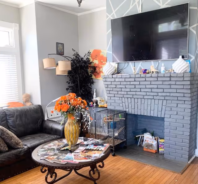 A cozy living room with a black leather couch, a round wooden coffee table with a vase of orange flowers and magazines, a gray brick fireplace with decorative items on the mantel, a large flat-screen TV mounted above the fireplace, a tall floor lamp with three beige lampshades, and a potted plant in the corner.