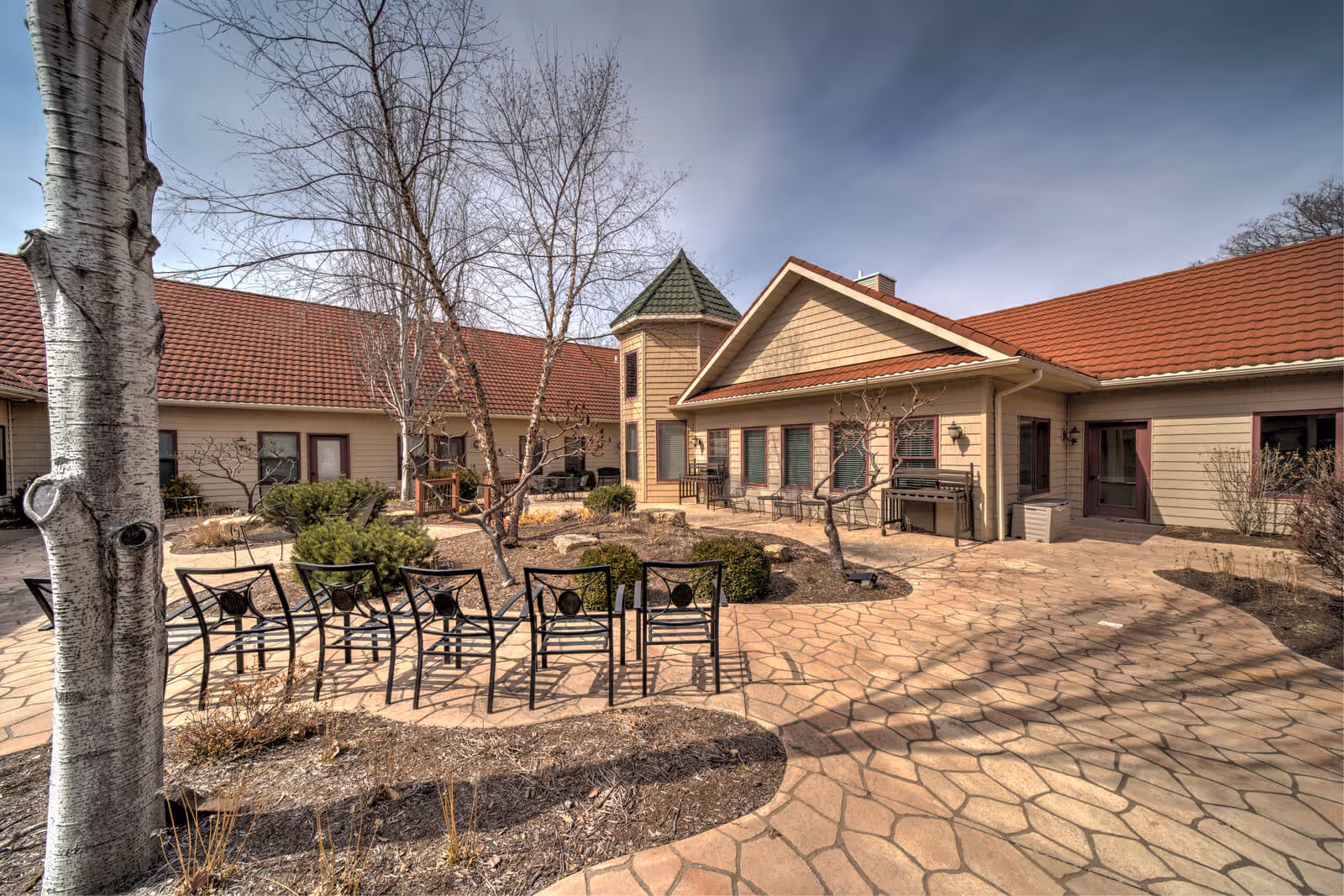 Outdoor courtyard area of a senior living facility with a paved stone patio, several metal chairs arranged in a row, leafless trees, shrubs, and a beige building with a red tiled roof surrounding the courtyard.