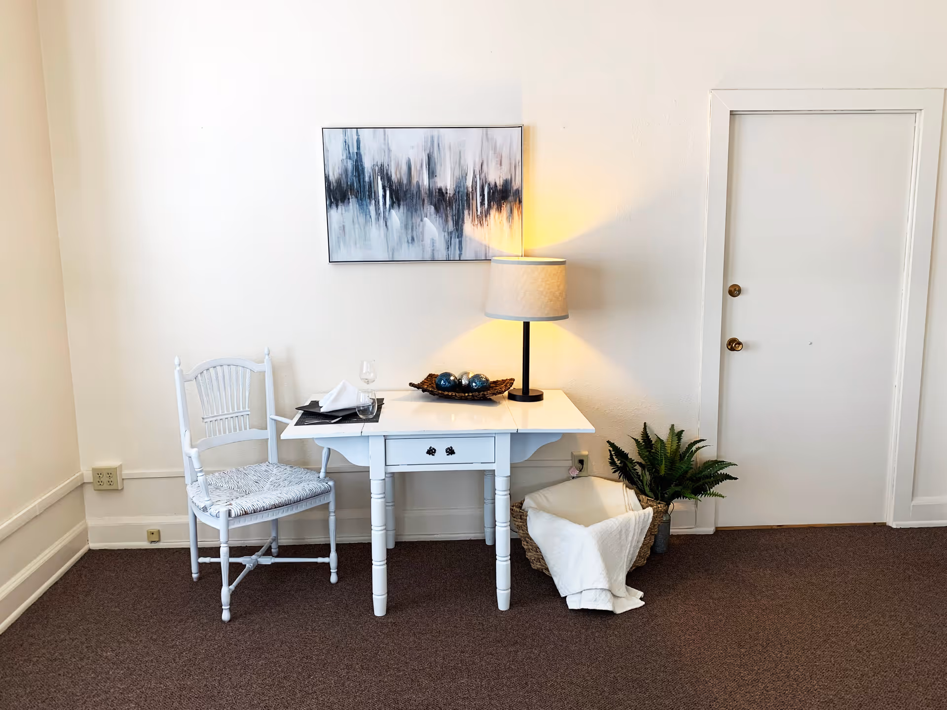 A small white wooden table with a drawer, set against a white wall. On the table are a lamp with a beige shade, a decorative basket with three blue and silver balls, a folded white napkin, a wine glass, and a drinking glass. To the left of the table is a white wooden chair with a woven seat. To the right of the table is a basket with a white blanket and a green potted plant. Above the table hangs an abstract painting with black, white, and gray tones. A closed white door with two brass knobs is visible on the right side of the image. The floor is carpeted in brown.