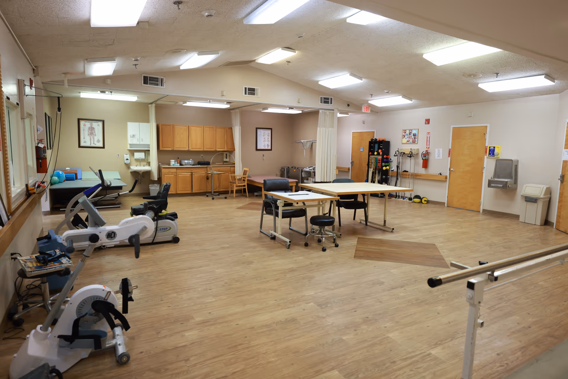 A spacious rehabilitation room with exercise equipment including stationary bikes, therapy tables, chairs, and parallel bars. The room has wooden flooring, bright fluorescent lighting, cabinets, and various therapy tools and equipment along the walls.
