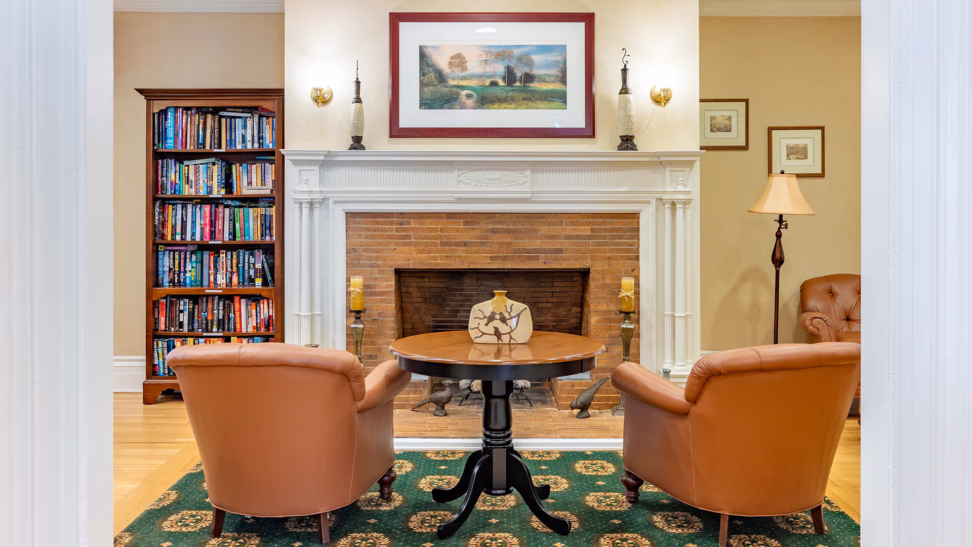 A cozy living room area with two brown leather armchairs facing a brick fireplace with a white mantel. A round wooden table with a decorative vase is placed between the chairs on a green patterned rug. To the left, there is a tall bookshelf filled with books, and to the right, a floor lamp and framed pictures on the wall.