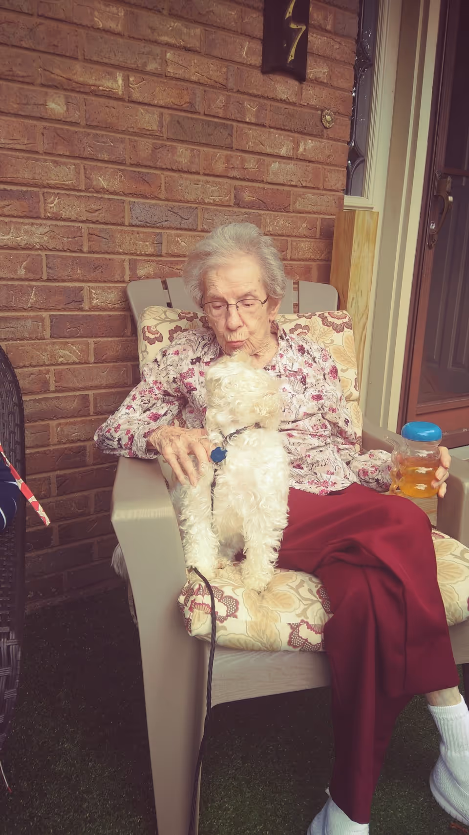 An elderly woman with glasses sitting on a cushioned chair outside against a brick wall, holding a small white dog on her lap and a plastic cup with a yellow drink in her right hand.