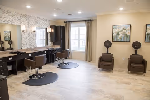 A bright and spacious hair salon area with two styling chairs in front of large mirrors and two hair washing stations with brown chairs. The room has beige tiled floors, cream-colored walls, a large window with beige curtains, and two framed floral paintings on the wall.