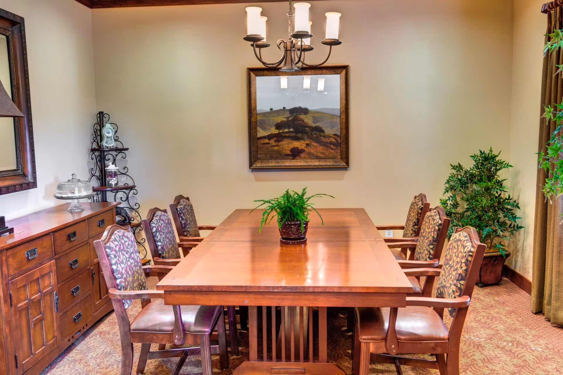 A dining room with a long wooden table surrounded by eight upholstered chairs with floral patterns. A green potted plant is placed in the center of the table. On the left side, there is a wooden sideboard with a glass cake stand and decorative items. A framed landscape painting hangs on the beige wall above the table, and a chandelier with five lights is suspended from the ceiling. There are two large potted plants in the corners of the room, and beige curtains hang on the right side.