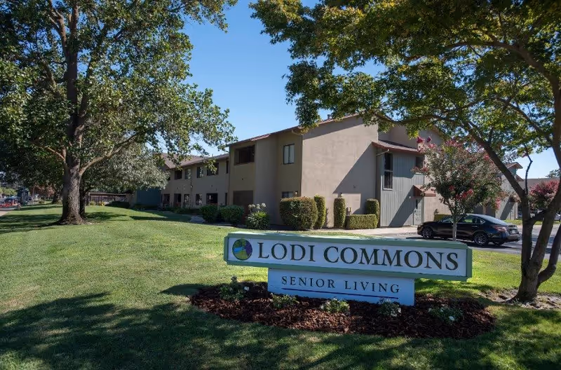 Exterior view of Lodi Commons Senior Living facility with a large green lawn, trees, and a sign in the foreground that reads 'Lodi Commons Senior Living'. The building is two stories with beige and gray walls and several windows. A car is parked near the building.