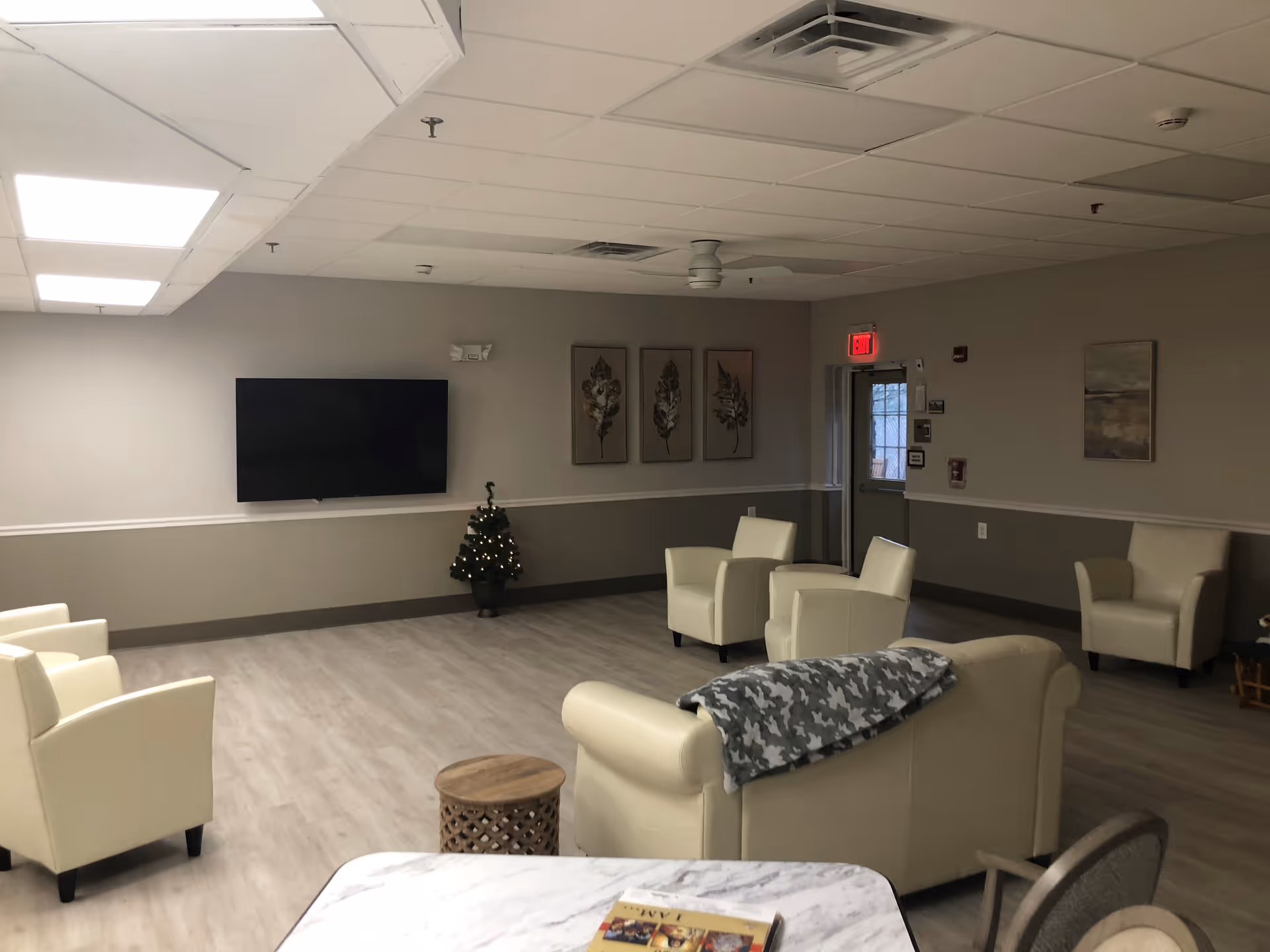A spacious living room area with beige walls and light wood flooring. The room contains several cream-colored armchairs and a matching sofa with a gray patterned throw draped over it. A small round wooden side table is in front of the sofa. On the wall, there is a large flat-screen TV mounted above a small decorated Christmas tree with lights. Three framed leaf artworks hang on the wall next to an exit door with a red exit sign above it. The ceiling has recessed lighting and a ceiling fan.