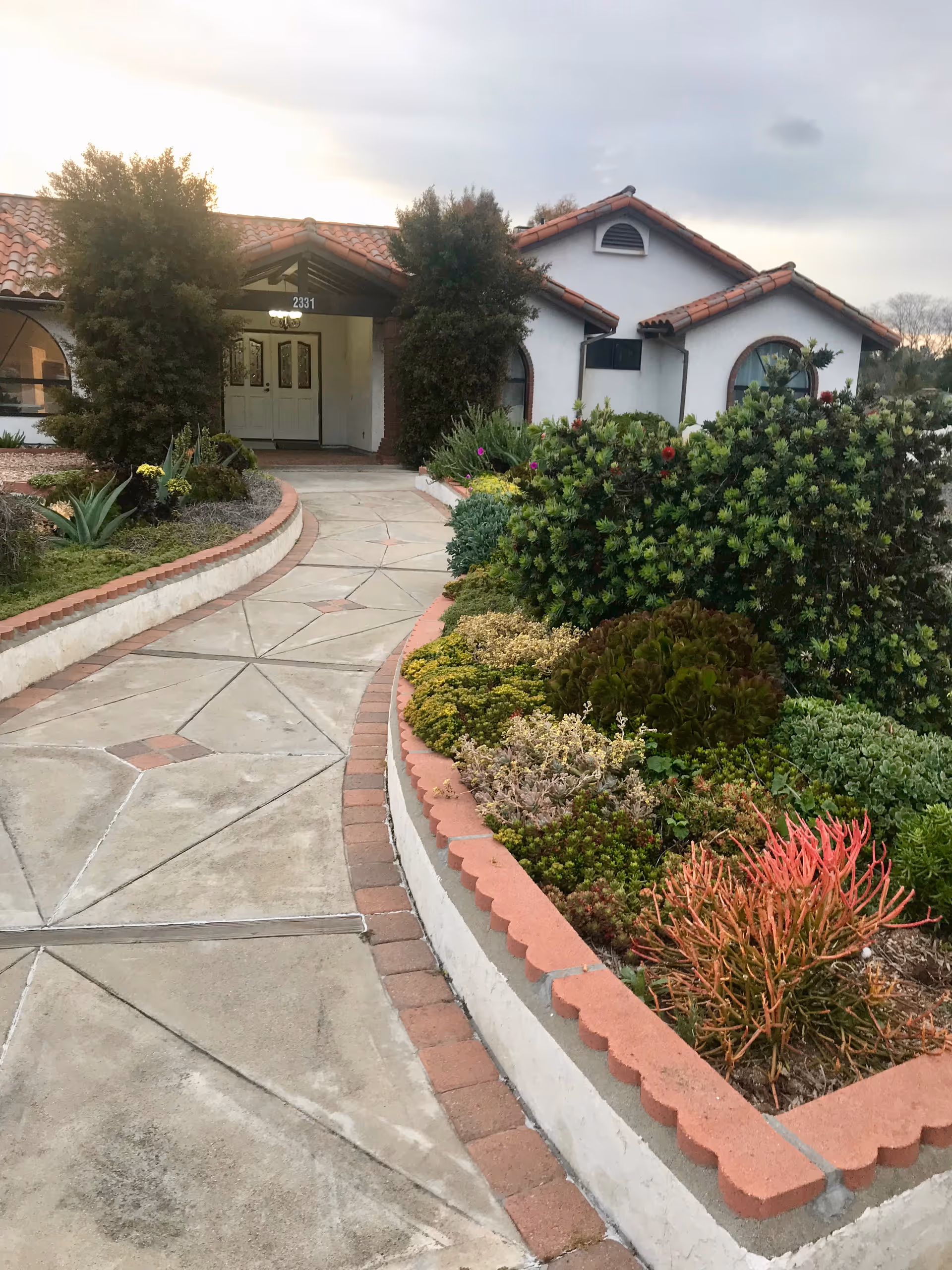 A paved walkway with geometric patterns leads to the entrance of a single-story building with white walls and a red-tiled roof. The walkway is bordered by a raised garden bed filled with various green and red succulent plants and shrubs. Two tall bushes flank the entrance door, and the building number 2331 is visible above the door. The sky is overcast.