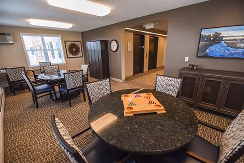 Interior view of a common area in a senior living facility with a round table in the foreground featuring a board game, surrounded by chairs. In the background, there are additional tables and chairs near a window, a wall clock, a cabinet, and a painting on the wall.