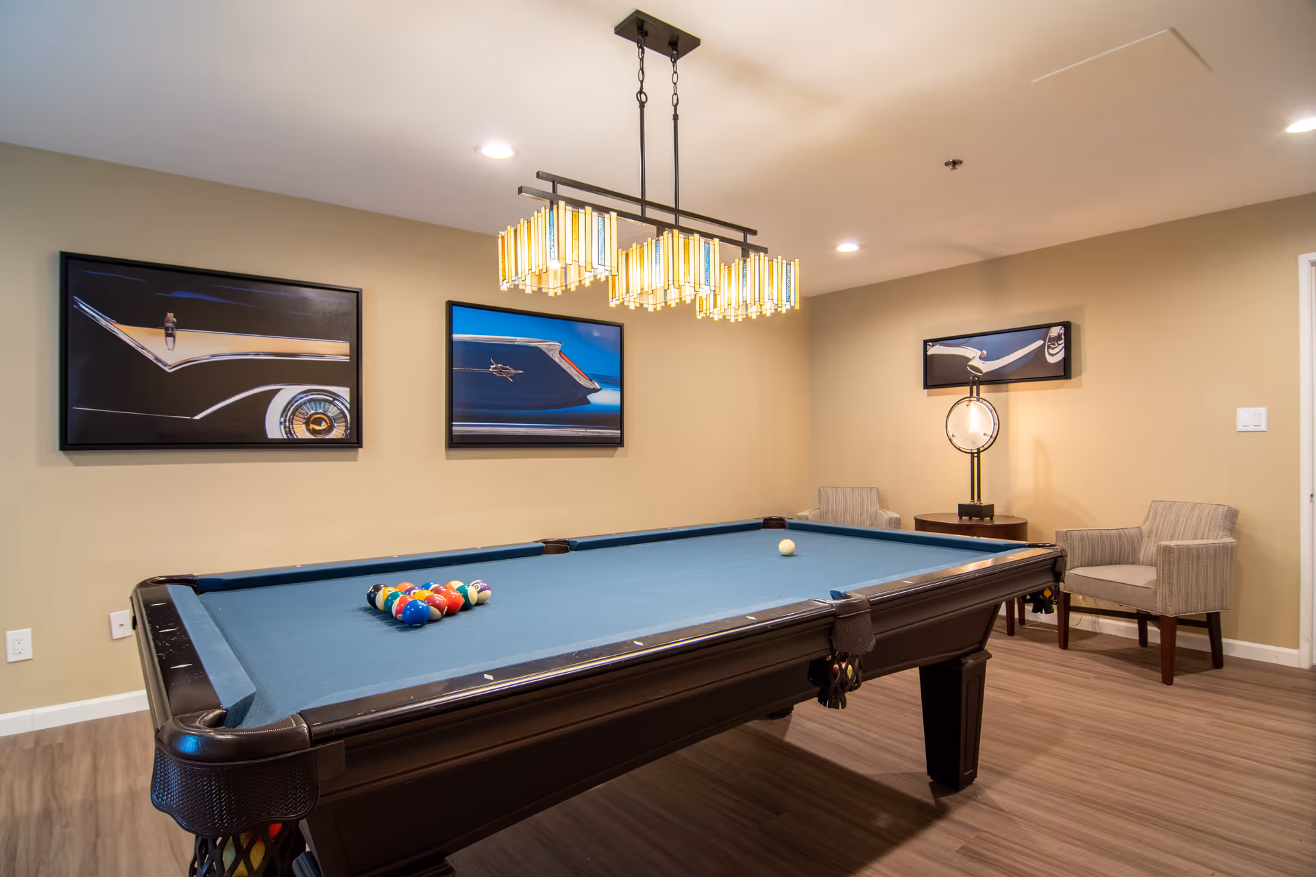 A recreation room with a pool table set up for a game, featuring a blue felt surface and a rack of colorful billiard balls. The room has beige walls adorned with framed pictures of classic car details. There are two upholstered armchairs and a small round table with a decorative lamp in the corner. A modern rectangular chandelier hangs above the pool table, and the floor is covered with wood-style flooring.