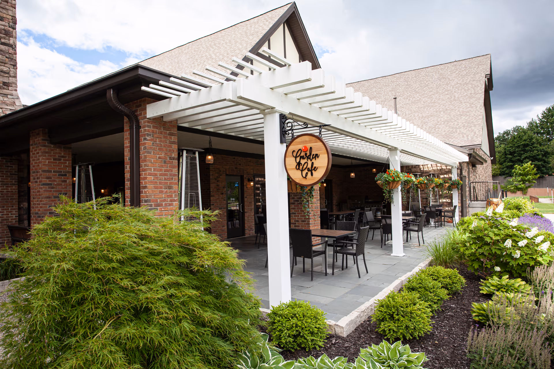 Outdoor patio area of a brick building with a white pergola overhead. Several tables and chairs are arranged under the pergola, with hanging flower baskets attached to the pergola beams. A round wooden sign reading 'Garden Cafe' is hanging from one of the pergola posts. The patio is surrounded by well-maintained greenery and flowering plants, with a partly cloudy sky in the background.