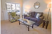 A cozy living room with a gray sofa adorned with patterned pillows, a floral armchair with a green cushion, a glass coffee table, two side tables with lamps, a decorative mirror on the wall, and a window letting in natural light.