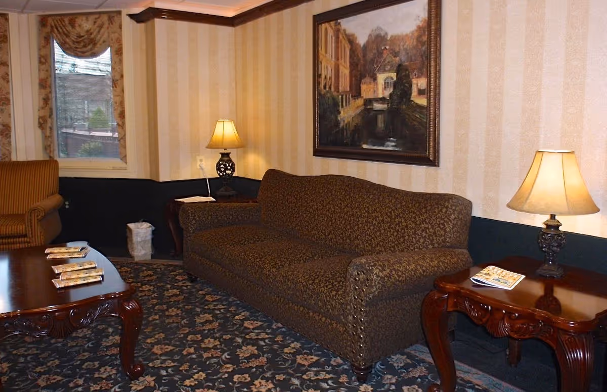 A cozy living room with a patterned brown sofa and a matching armchair. There are two wooden side tables with decorative lamps on each side of the sofa. A wooden coffee table with some magazines is in front of the sofa. The walls have striped wallpaper and a framed painting depicting a canal scene. A window with floral curtains is visible in the background.