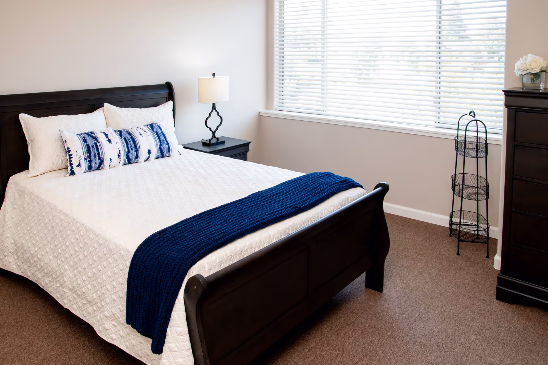 A neatly made bed with a dark wooden frame, white quilted bedding, and decorative blue and white pillows. A dark wooden nightstand with a lamp is beside the bed. A window with white blinds allows natural light into the room. There is a dark wooden dresser with a vase of white flowers on top and a black metal three-tiered stand in the corner of the room.