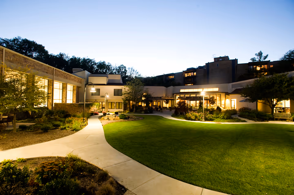 Evening view of a senior living facility courtyard with well-maintained green lawn, curved concrete pathways, outdoor lighting, and surrounding buildings with large windows illuminated from inside.