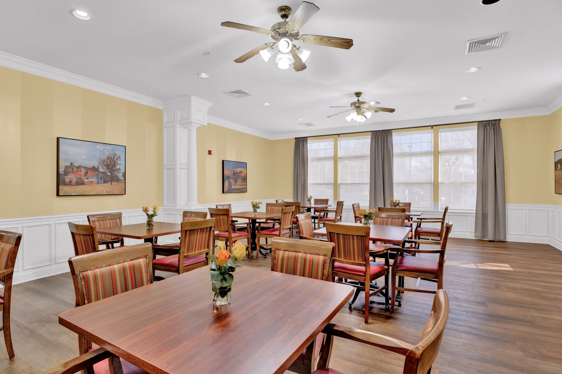 A bright dining room with multiple wooden tables and chairs arranged neatly. Each table has a small vase with flowers. The room has yellow walls with white wainscoting, large windows with gray curtains, ceiling fans with lights, and framed paintings on the walls.