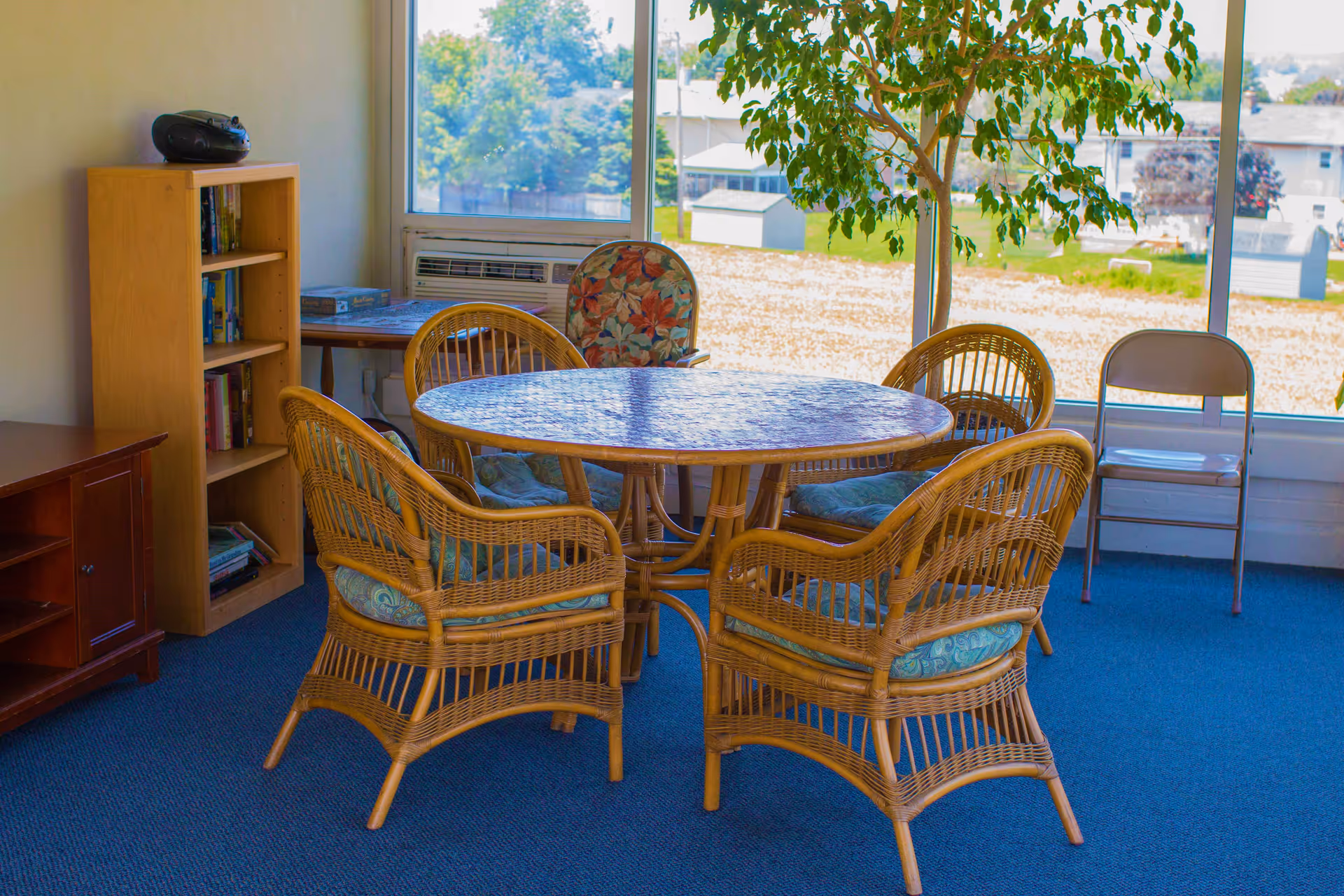 A sunlit communal room with a round wicker table and matching wicker chairs by large windows.