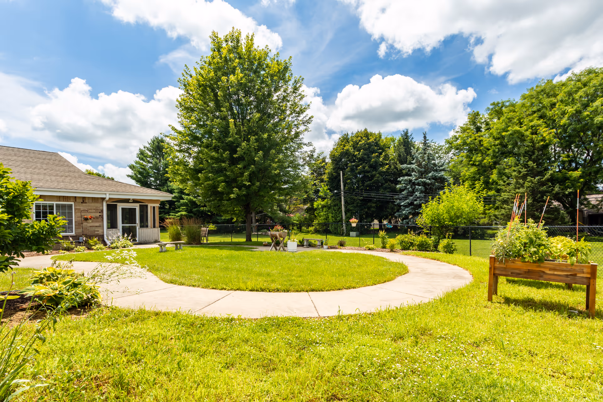 Sunlit circular courtyard with a grassy lawn, paved walkway, trees, and a single-story facility building at left.