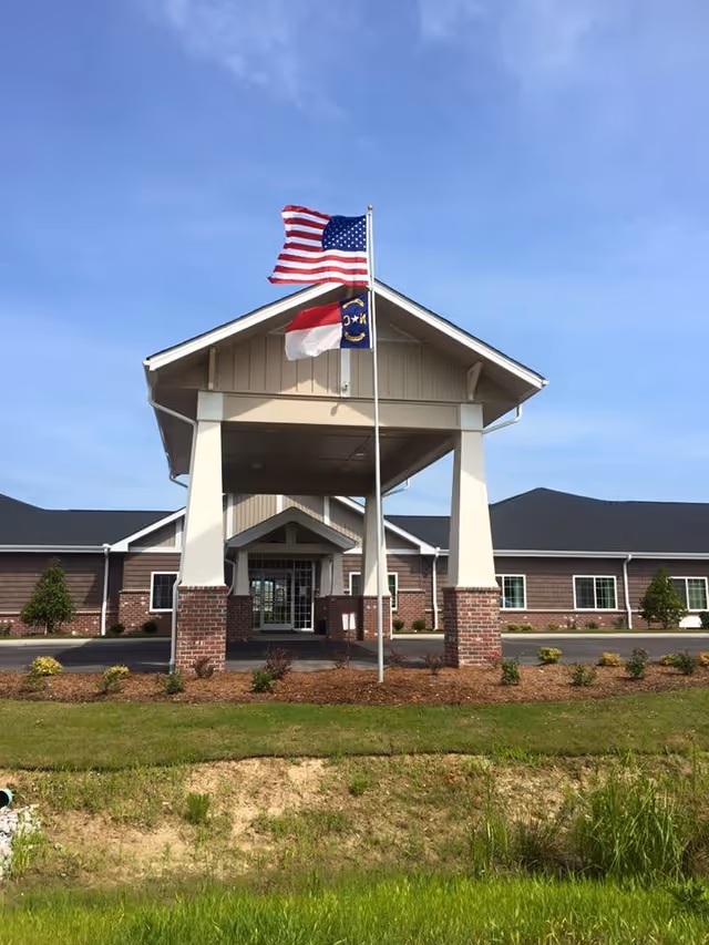 Front exterior view of a single-story building with a covered entrance supported by white columns and brick bases. Two flags, the American flag and a state flag, are flying on a flagpole in front of the entrance. The building has a dark roof and light-colored siding with several windows. The foreground shows a grassy area and some landscaping.