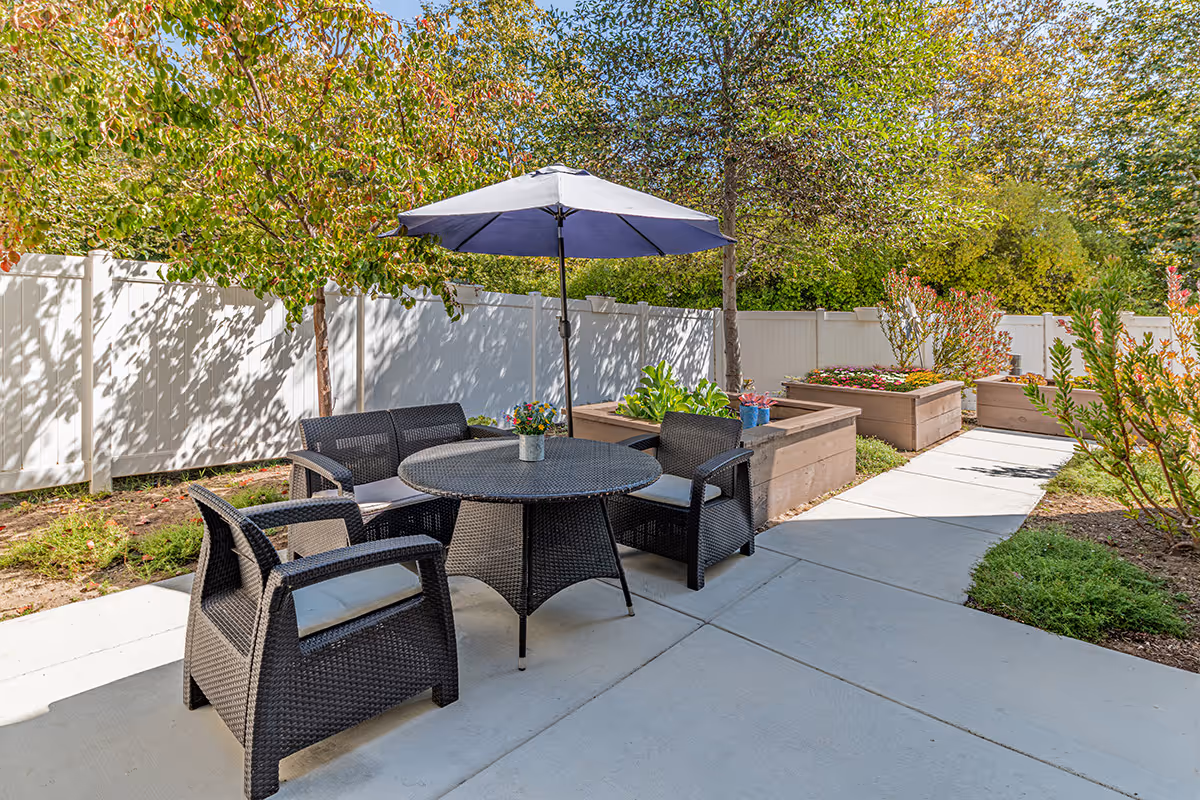 Patio seating area with a round table, four wicker chairs and an umbrella next to raised planter boxes and a white fence.