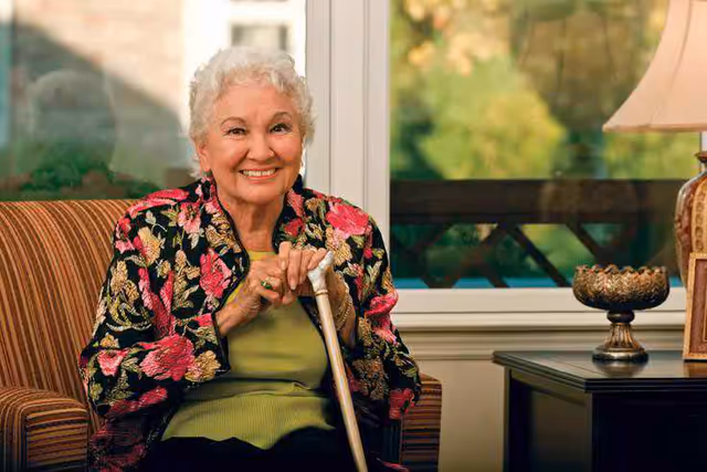 An elderly woman with short white hair sitting on a striped armchair indoors, smiling and holding a wooden cane. She is wearing a floral patterned jacket over a green top. Behind her is a window showing greenery outside and a table with a decorative bowl and a lamp.
