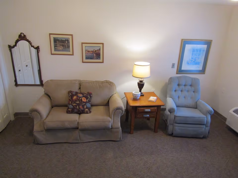 Small living room with a beige loveseat, patterned throw pillow, wooden side table with a lamp, and a blue upholstered recliner under framed artwork.