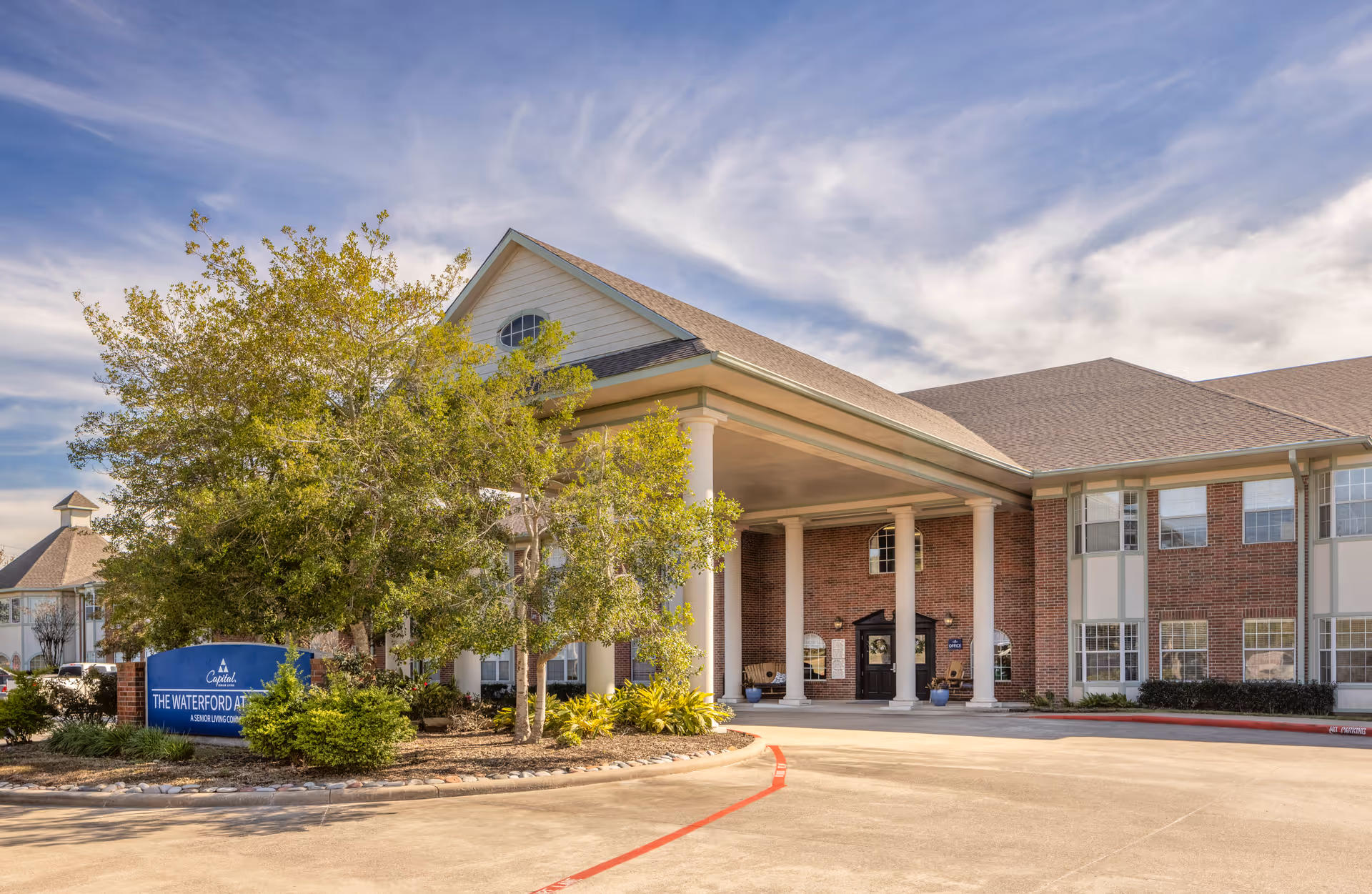 Exterior view of The Waterford at Baytown senior living community building with a covered entrance supported by white columns, surrounded by trees and landscaping under a partly cloudy sky.
