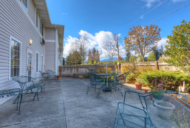 Outdoor patio with metal tables and chairs next to a light-colored building and a fenced garden under a blue sky.