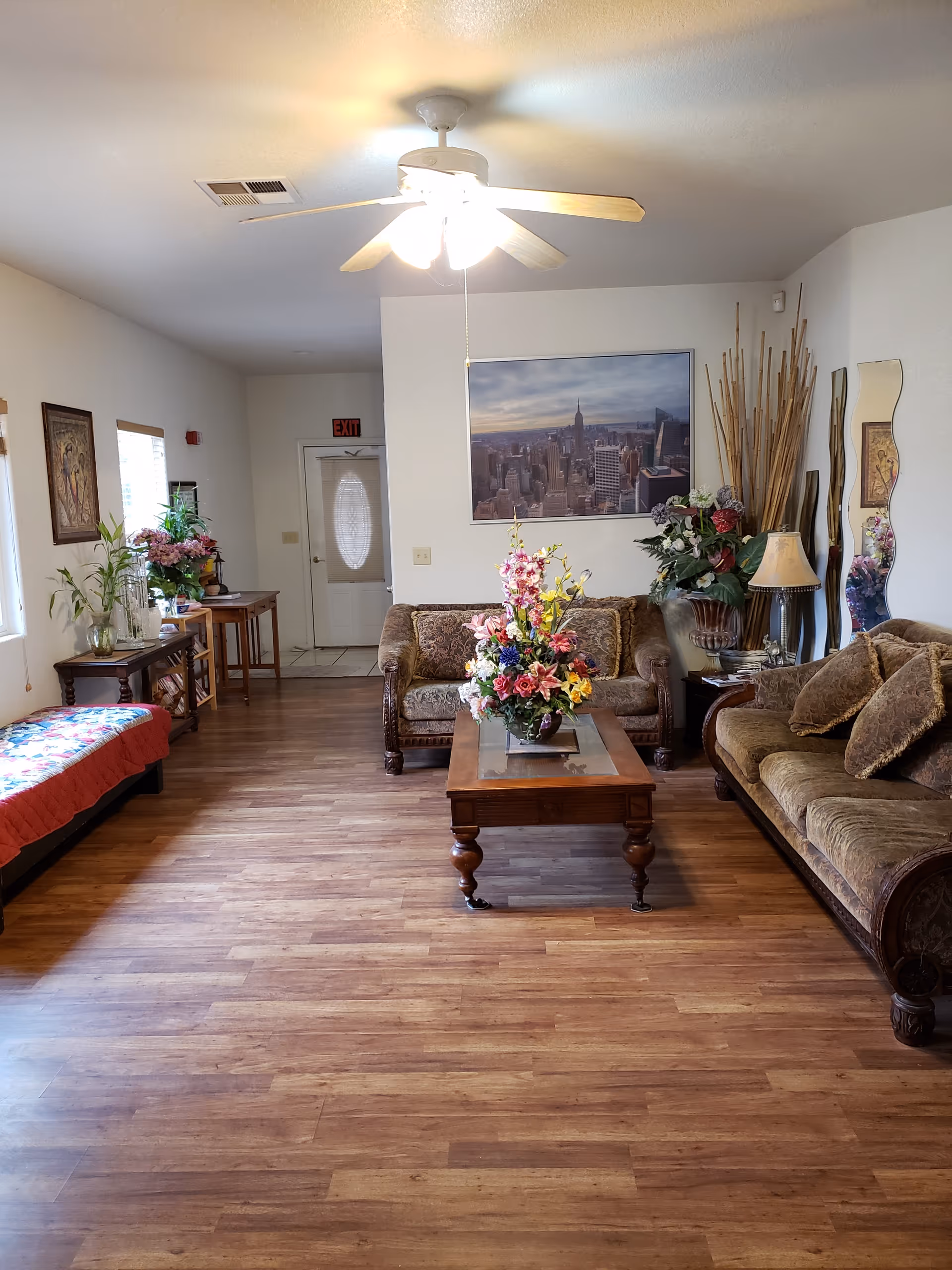 A cozy living room with wooden flooring, two brown upholstered sofas with cushions, a wooden coffee table with a floral arrangement, a ceiling fan with lights, a large framed cityscape photo on the wall, decorative plants, a lamp, and a mirror. There is a bench with a colorful cushion near the windows on the left side and a small table with more plants near the entrance door in the background.
